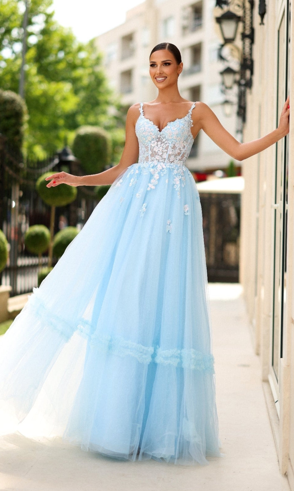 A woman stands outdoors, smiling with one hand on a wall, wearing the Tina Holly TY405 long formal dress in pastel blue, featuring a corset bodice with floral embroidery and a flowing tulle skirt.